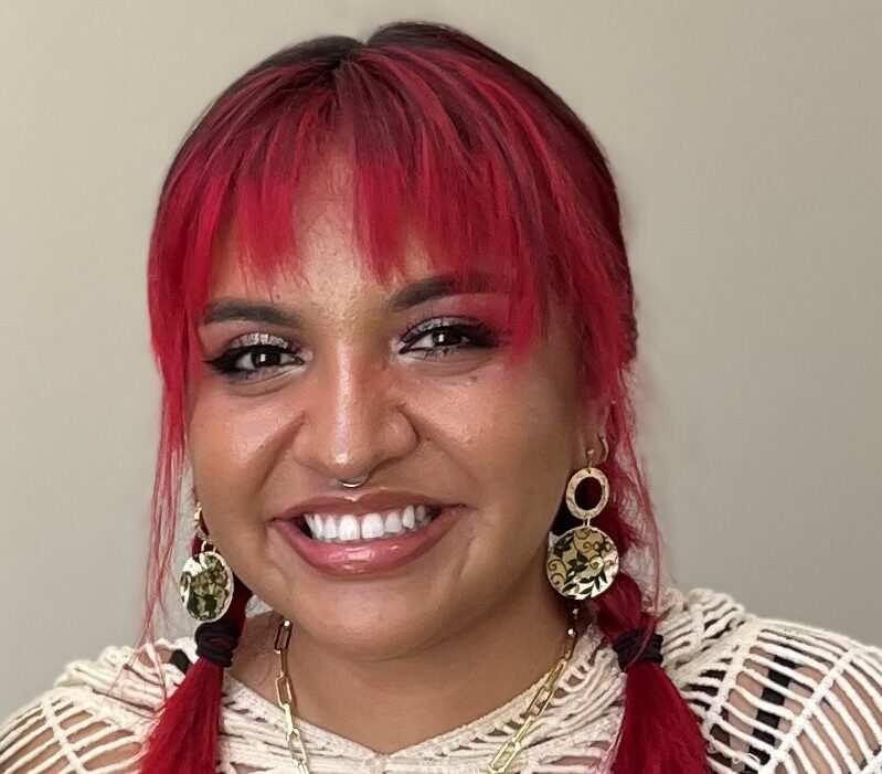 A photo of a smiling Latina with large earrings, a cream-colored sweater, and vivid red hair worn in braided pigtails.