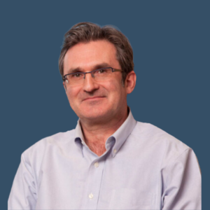 A man with short, graying hair, wearing glasses and a light blue button-up shirt. He is smiling gently and posed in front of a solid dark blue background.