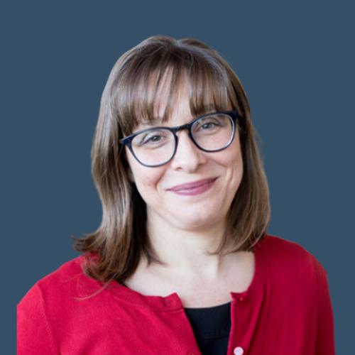 A professional headshot of a woman with shoulder-length brown hair, bangs, and wearing glasses. She has a gentle smile and is dressed in a red cardigan over a black top. The background is a solid, muted blue color.