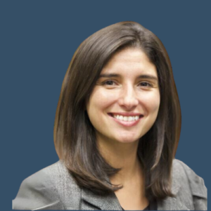 A headshot of a woman with shoulder-length dark hair, smiling. She is wearing a gray blazer, set against a blue background.