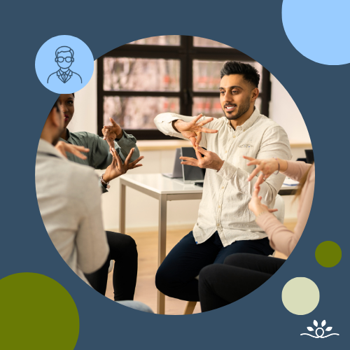 A man seated in a bright classroom with large windows, leading a small group discussion in American Sign Language. His hands are positioned mid-sign, and other participants’ hands are partially visible in the frame. The circular crop is set against a dark blue background with green, cream, and blue accent circles. Upper left has a white faculty icon in a blue circle. NDCSS logo bottom right.