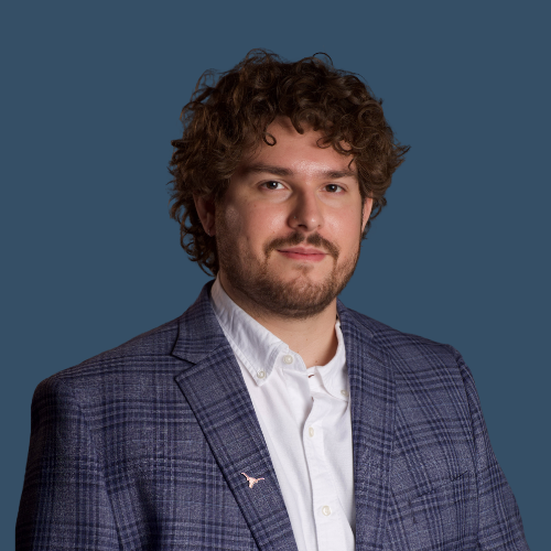 A professional headshot of a man with curly brown hair and a beard. He is wearing a blue plaid blazer with a white dress shirt underneath. The man is facing forward with a slight smile. The background is a solid, muted blue color.