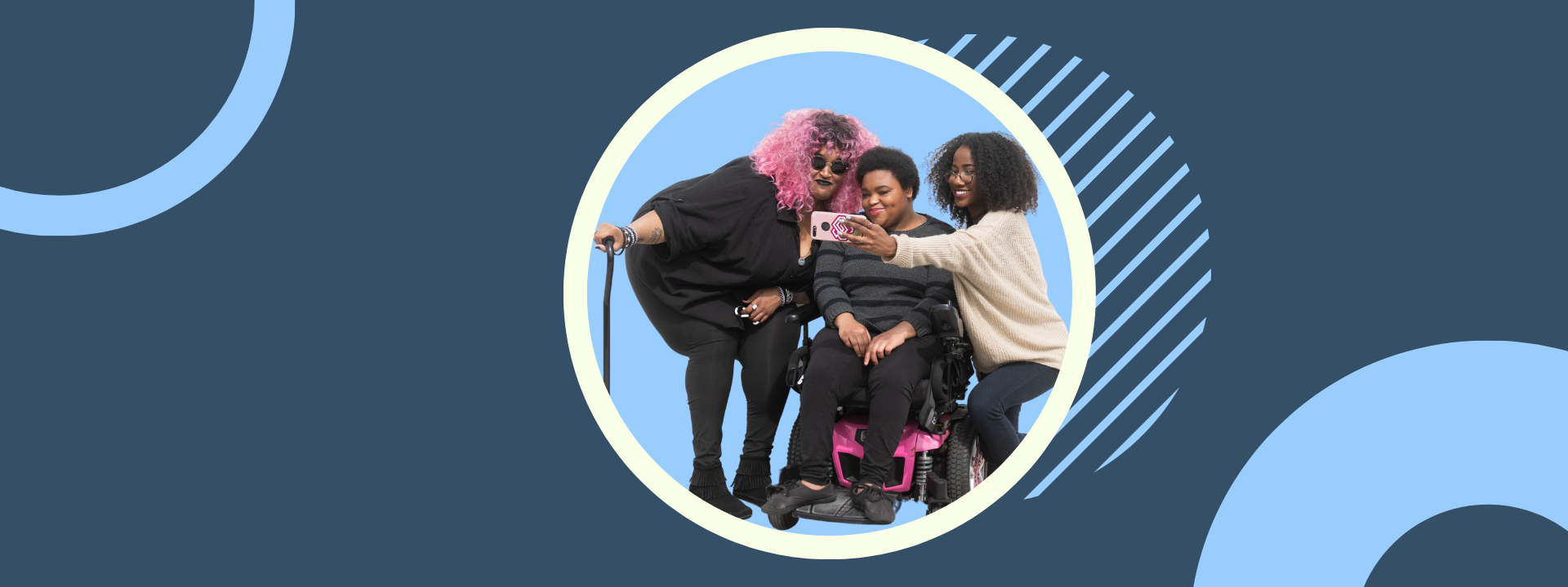 A zoomed out full body shot showing three Black and disabled friends (a non-binary person with a cane and tangle stim toy, a non-binary person sitting in a power wheelchair, and an invisibly disabled femme) smiling and taking a cell phone selfie together. All are outdoors in front of a white wall.
