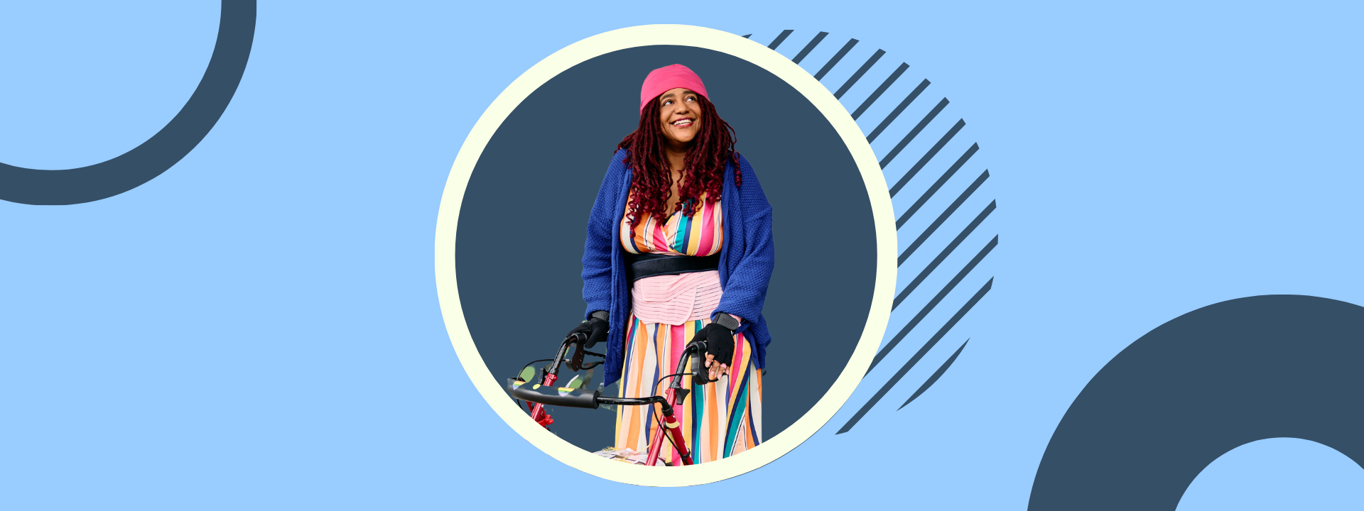 A non-binary Black woman stands in front of a shop with their sticker-covered rollator, smiling and looking off in the distance. Their colorful outfit includes a beanie hat, cardigan, waist trainer filled with ice packs, rainbow-striped dress, bike gloves layered over arthritis gloves, rainbow heart leggings, and sneakers.