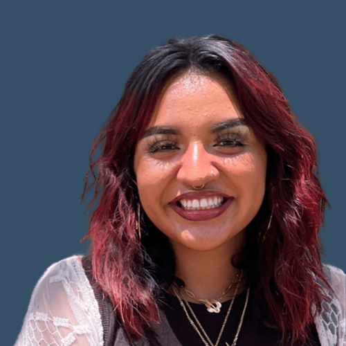 A woman with shoulder-length, wavy black and red hair. She is smiling brightly and has a nose ring. She is wearing a black shirt with a lacy white cover-up and layered necklaces. The background is solid dark blue.