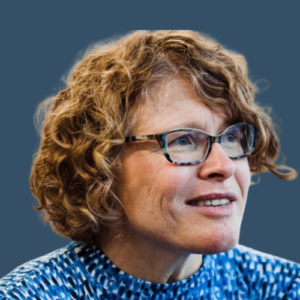 A headshot of a woman with curly light brown hair, wearing glasses and a blue patterned top. She is smiling and looking to the side, set against a blue background.