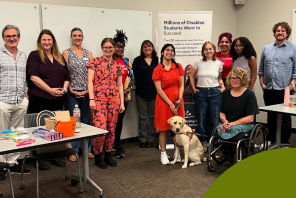 A diverse group of 13 people and one guide dog are in a college classroom, surrounding a vertical banner that reads “Millions of disabled students want to succeed.