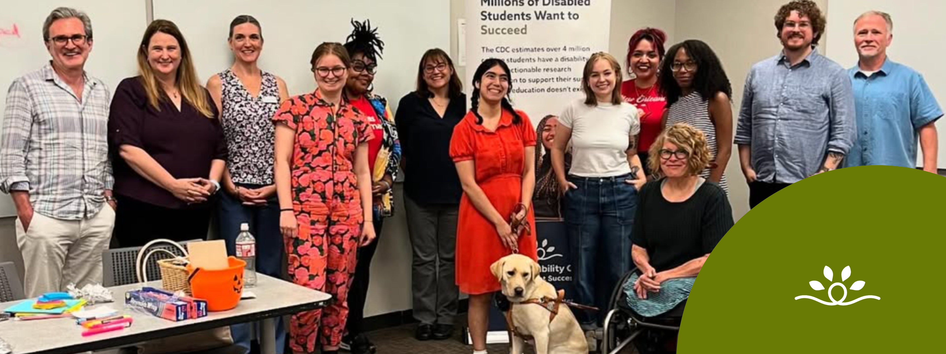 A diverse group of 13 people and one guide dog are in a college classroom, surrounding a vertical banner that reads “Millions of disabled students want to succeed.