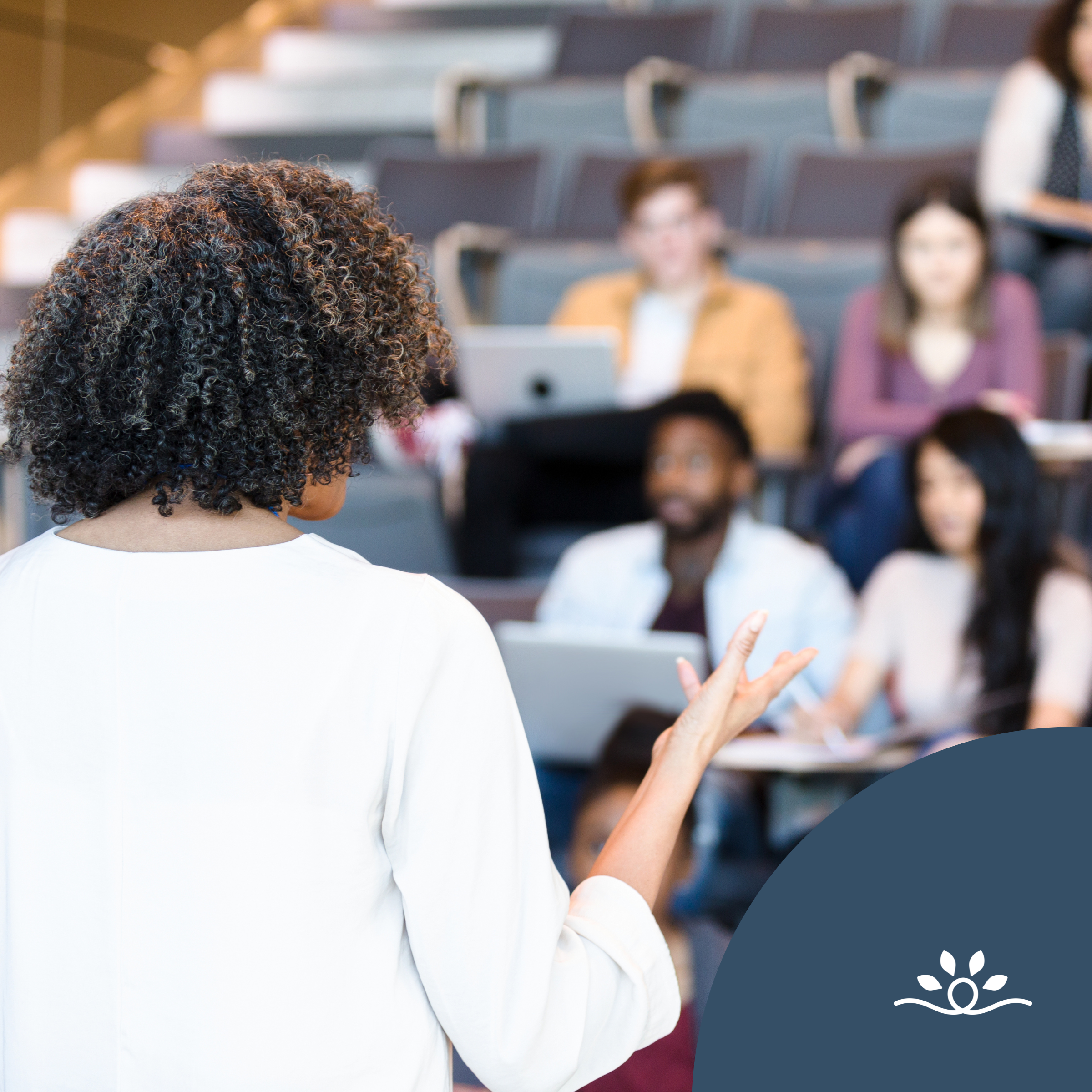 Black woman standing with her back to the camera wearing a white t-shirt with shoulder length brown coily hair that has lighter brown highlights. She is teaching a group of students that are blurred out to emphasize the professor practicing accessible teaching. Some students have a laptop. At the bottom left corner there is a blue semi circle with a national disability logo of a icon with a thick white line that loops in a circle in the middle and curves down and upward at each end. Above the line are four white spear-shaped leaves arranged in an arc over the loop.