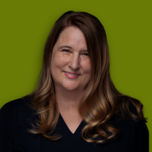 A headshot of Stephanie Cawthon with long, wavy, light brown hair. She is smiling and wearing a dark top, set against a green background.
