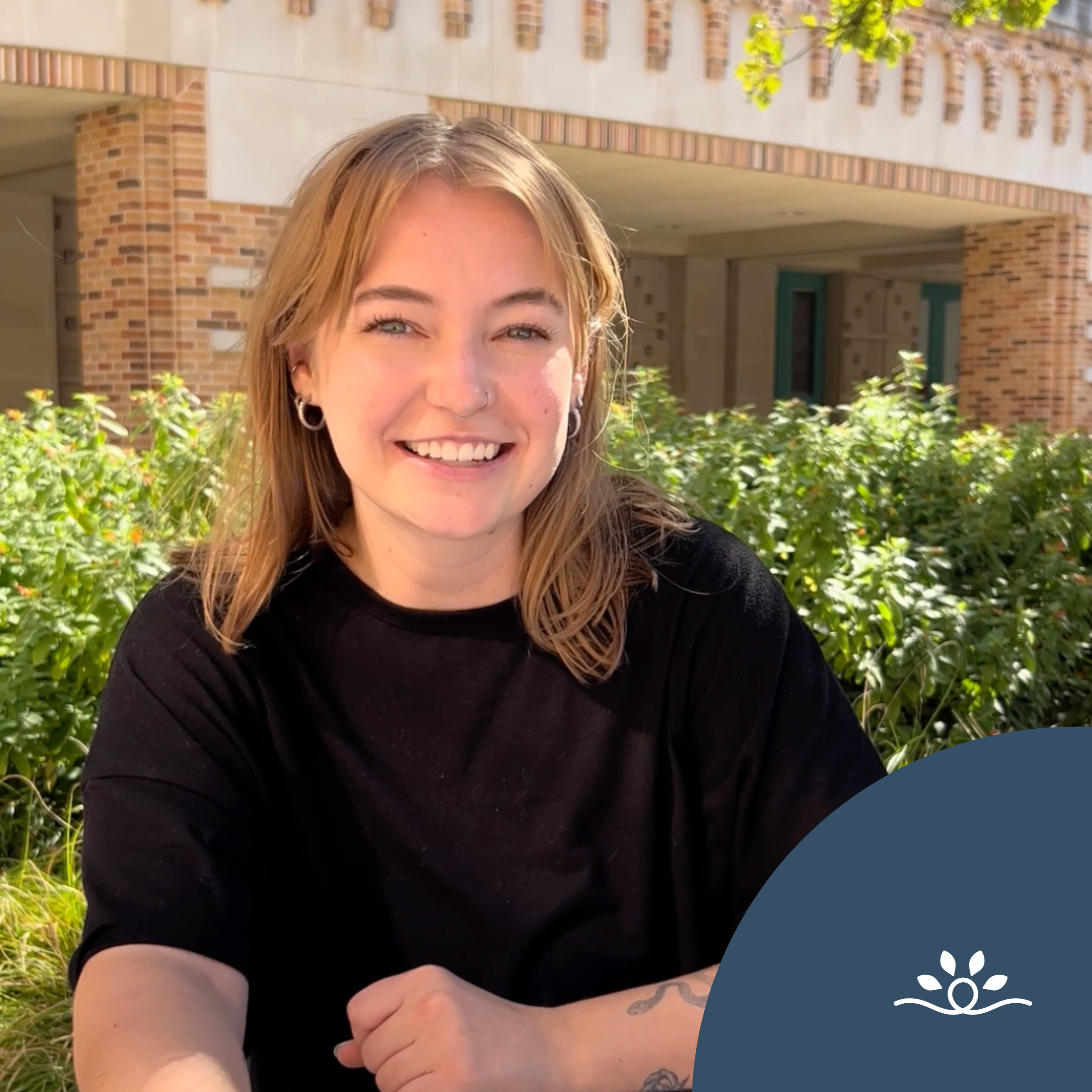 This graphic features a smiling person with light hair seated outdoors in front of lush green foliage and a building with brick and beige accents. The person is wearing a black top, and their arms are positioned casually in front of them. The background shows part of the building's exterior, including rectangular windows and an archway design at the top. In the bottom right corner, there is a dark blue overlay featuring the National Disability Center logo in white, which consists of a circular loop with four spear-shaped leaves arranged above it.