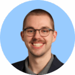 A man with short-cropped light brown hair, a trimmed mustache, and glasses smiles widely. He is wearing a gray collared shirt and a dark blazer. The background is a soft blue circle. Headshot for Binghamton University.
