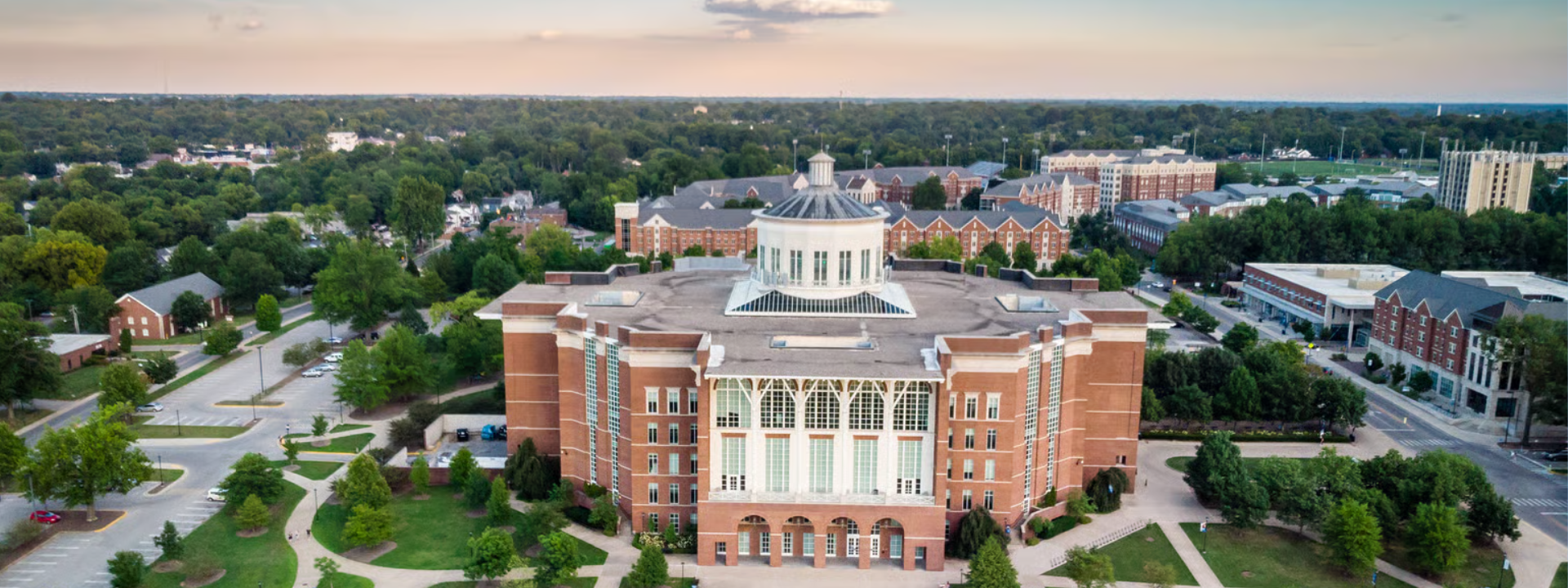 Aerial view of a large red-brick campus building with a white rotunda roof at its center, surrounded by trees, walking paths, and parking lots. The image captures a wide landscape of the University of Kentucky campus at sunset, with additional academic and residential buildings visible in the background. The layout includes symmetrical green spaces and a broad view of the surrounding cityscape.