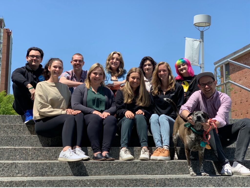 A group of ten people and one dog sit together on outdoor campus steps at Binghamton University under a clear blue sky. The group includes a mix of men and women, casually dressed, smiling at the camera. A man on the far right kneels next to a brown-and-white dog wearing a green bandana and holding a rope toy. A flagpole and red-brick buildings are visible in the background.