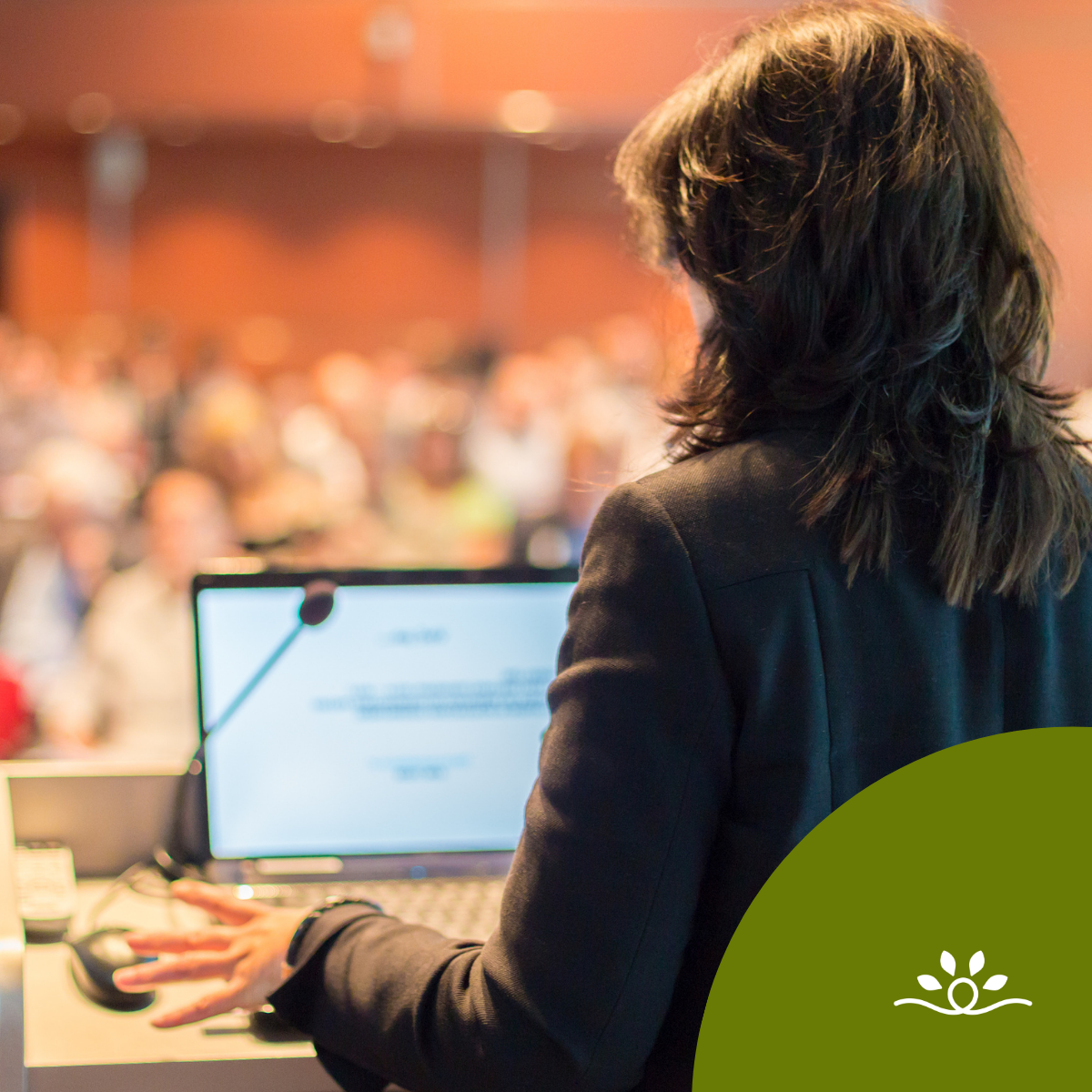 A blurred background of an auditorium filled with people in warm lighting. In the foreground, a person with dark shoulder-length hair is seen from behind, standing at a podium with a microphone and an open laptop. A dark olive-green circular shape with the white National Disability Center logo overlays the bottom right corner.