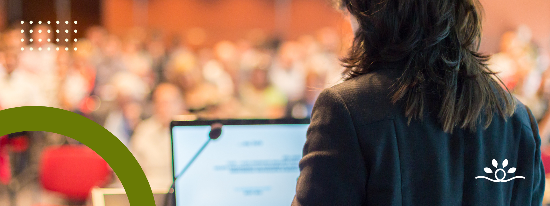 A wide horizontal design showing a blurred auditorium of people with warm tones in the background. In the foreground, the back of a person with dark shoulder-length hair at a podium with a microphone and laptop. A dark olive-green arc curves from the bottom left, while dotted white graphics appear near the top left. The white National Disability Center logo is placed on the lower right.