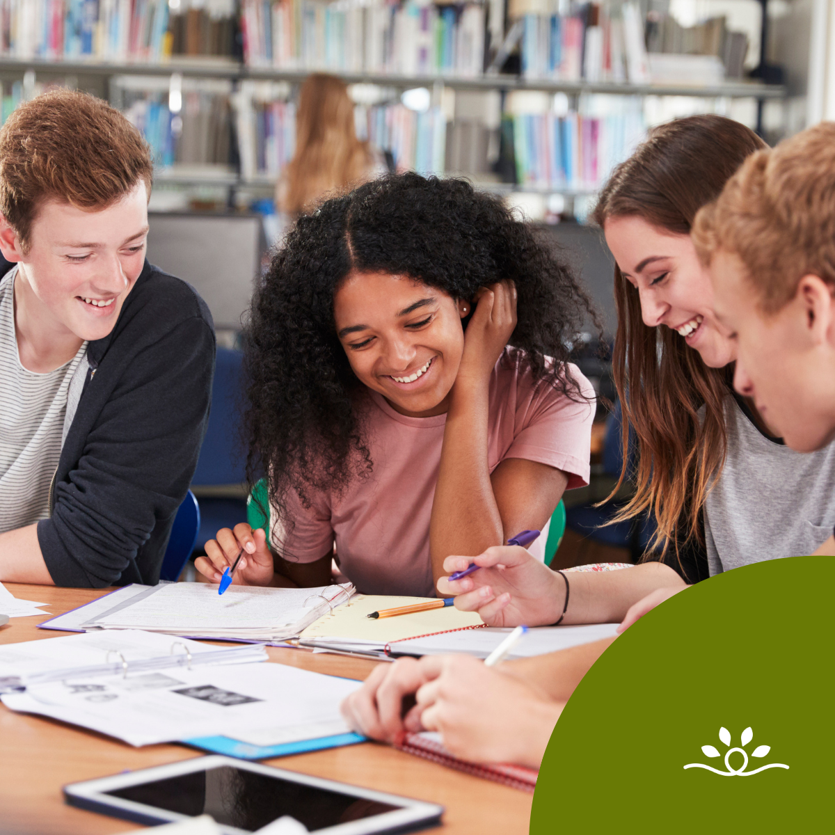 Four students sit together at a library table with books and notes spread out, smiling and writing as they collaborate. Shelves of books create a blurred background. A dark olive-green circular shape with the white National Disability Center logo overlays the bottom right corner.
