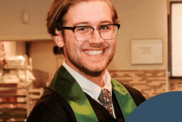 A close-up graduation portrait of a smiling Dalton Kendrick with light brown hair parted to the side, wearing black glasses, a nose ring, and a black gown with a green stole. He has a patterned tie and are standing indoors, with a softly blurred background of a room featuring warm lighting and framed wall decor. In the lower right corner, a dark blue circular overlay with a small white National Disability Center logo featuring four upward leaves above a curved line is visible.