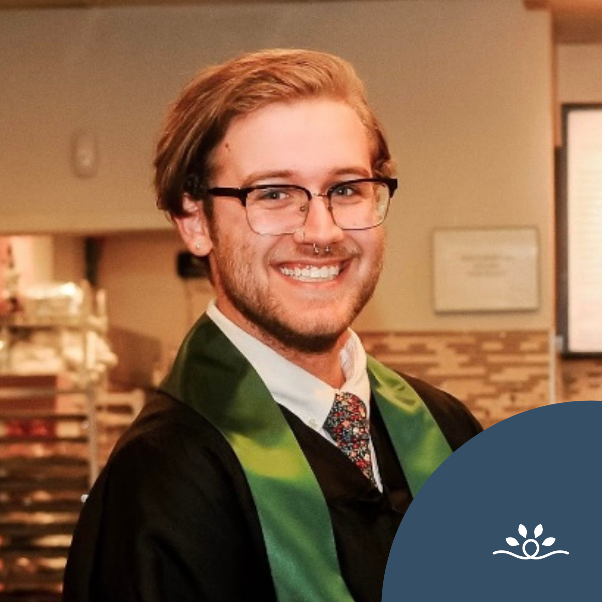 A close-up graduation portrait of a smiling Dalton Kendrick with light brown hair parted to the side, wearing black glasses, a nose ring, and a black gown with a green stole. He has a patterned tie and are standing indoors, with a softly blurred background of a room featuring warm lighting and framed wall decor. In the lower right corner, a dark blue circular overlay with a small white National Disability Center logo featuring four upward leaves above a curved line is visible.