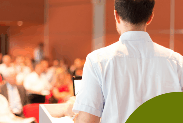 A male professor with short dark hair, wearing a light blue button-up shirt, stands at a podium with a laptop and papers while addressing an audience in a lecture hall. The audience appears slightly blurred in the background, seated in rows of red chairs. A green circular shape with a white leaf-and-wave logo overlays the bottom right corner, creating a branded visual accent.