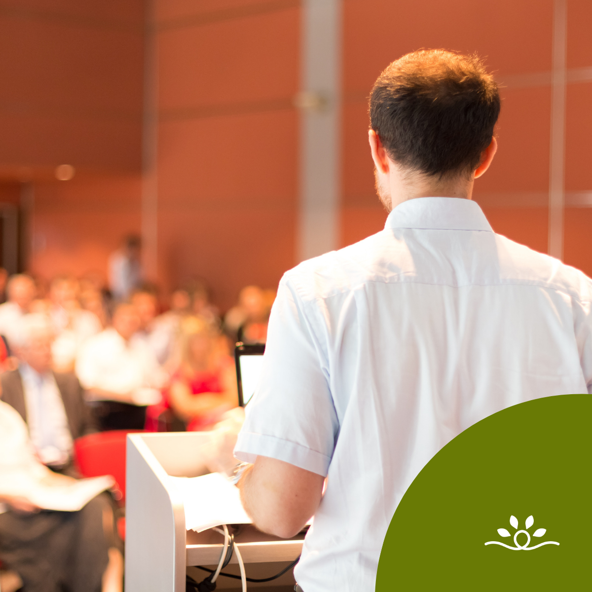 A male professor with short dark hair, wearing a light blue button-up shirt, stands at a podium with a laptop and papers while addressing an audience in a lecture hall. The audience appears slightly blurred in the background, seated in rows of red chairs. A green circular shape with a white leaf-and-wave logo overlays the bottom right corner, creating a branded visual accent.