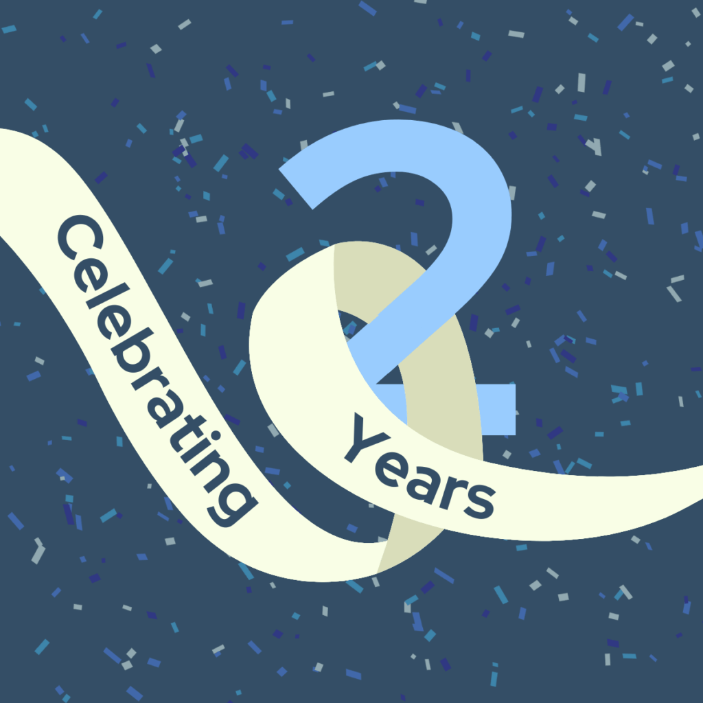 A dark blue background filled with scattered confetti pieces in shades of blue, purple, and gray. In the center, a large, light blue number “2” stands prominently. A cream-colored ribbon weaves around and through the number, with the words “Celebrating 2 Years” written in dark blue along the ribbon’s curve. The overall design conveys festivity and marks a two-year celebration.