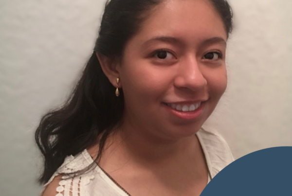 A head-and-shoulders portrait shows a young woman standing against a soft, light-colored background. She has long, dark hair pulled back, wears small gold drop earrings, and a sleeveless white top with delicate lace detailing along the shoulder. She smiles gently toward the camera. A curved dark blue graphic overlays the lower right corner, featuring the National Disability Center logo with a circular line and leaf motif.