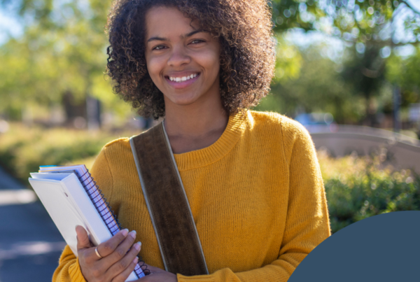 A college student stands outdoors on a campus walkway, smiling and holding notebooks. She wears a mustard yellow sweater and a shoulder bag, with trees and greenery in the background. The image represents student experiences in higher education, including decisions around disability disclosure and navigating access to support. A dark blue circular overlay with a white leaf-like logo appears in the bottom right corner.
