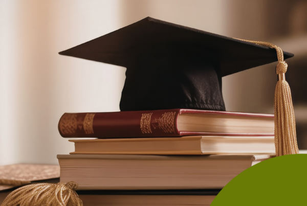 The image features a graduation cap placed atop a stack of books. The cap, made of black fabric, has a flat top and a long, golden tassel hanging from one side, signifying graduation. Below the cap, there are several books. The top book is maroon with decorative gold detailing. Beneath it, there are two additional books in lighter colors. The background is softly blurred.