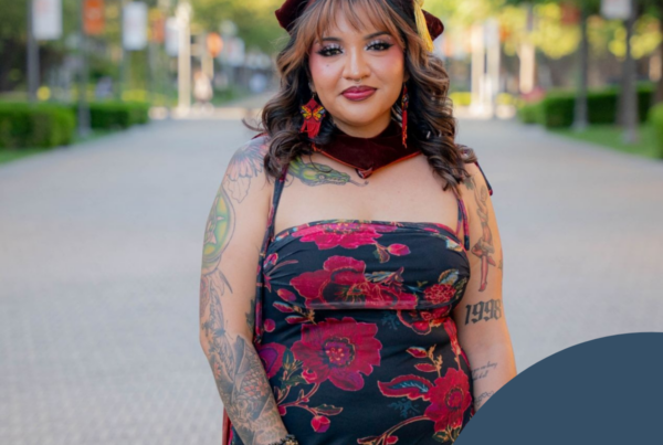 The image features a woman standing on a paved pathway lined with trees that extend into the distance. The individual is centered in the frame, wearing a black dress adorned with large red floral patterns. She has multiple tattoos on her arms, and her hands are decorated with rings and bracelets. The woman is also wearing a maroon cap atop their wavy hair and matching maroon earrings. The background showcases a clear day with lush green trees and blurred buildings visible in the far distance, creating a serene atmosphere.
