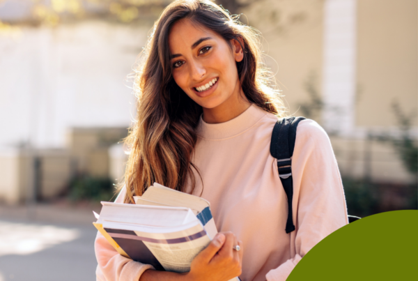 The image features a young woman standing outdoors, smiling warmly at the camera. She has long, wavy brown hair that cascades over her shoulders. She is dressed in a light pink sweatshirt, which contrasts with the darker elements of her outfit. In her arms, she carries several books stacked together, hinting at a student lifestyle. The background is softly blurred, showcasing hints of greenery and architecturally simple buildings, indicating an academic environment.