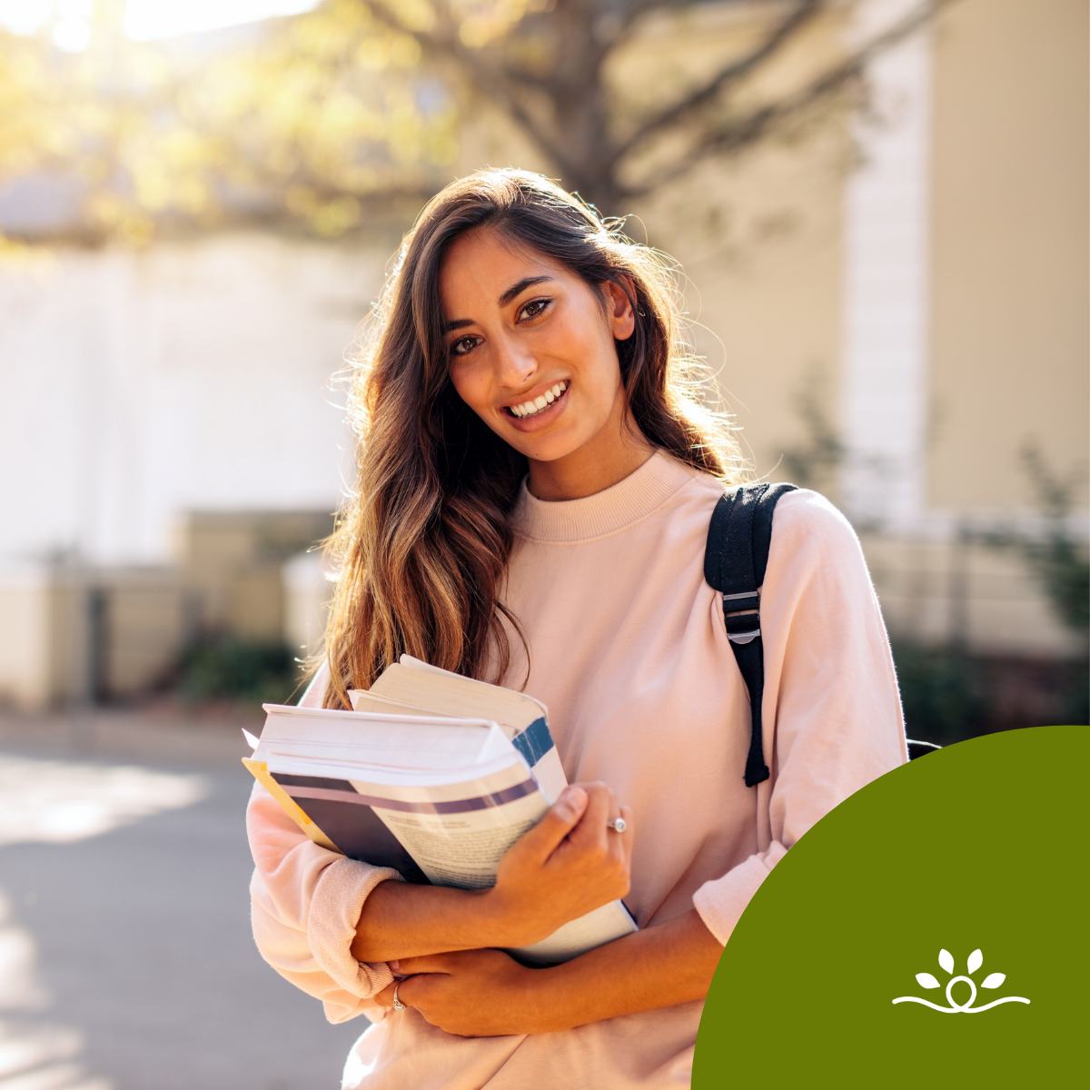 The image features a young woman standing outdoors, smiling warmly at the camera. She has long, wavy brown hair that cascades over her shoulders. She is dressed in a light pink sweatshirt, which contrasts with the darker elements of her outfit. In her arms, she carries several books stacked together, hinting at a student lifestyle. The background is softly blurred, showcasing hints of greenery and architecturally simple buildings, indicating an academic environment.