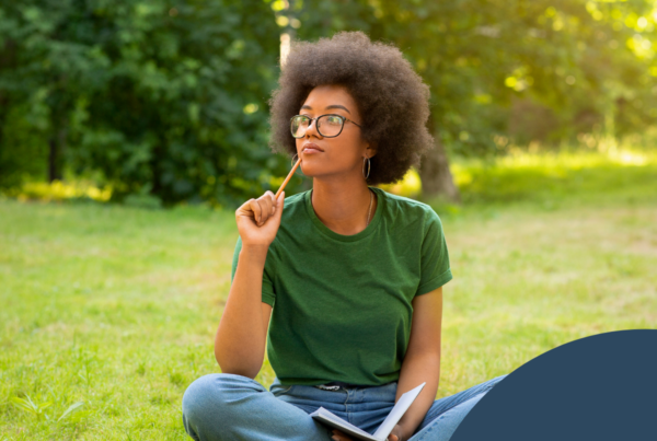 The image depicts a young woman sitting cross-legged on a grassy patch in a park. She has a full, curly afro hairstyle and wears large, round glasses. Dressed in a green t-shirt and light blue jeans, she appears to be deep in thought, holding a pencil to her chin. In her other hand, she holds an open notebook, suggesting she is engaged in some form of writing or contemplation. A small, disposable coffee cup is placed on the ground beside her. The background features lush greenery, with soft sunlight filtering through the trees, creating a serene and inviting atmosphere.