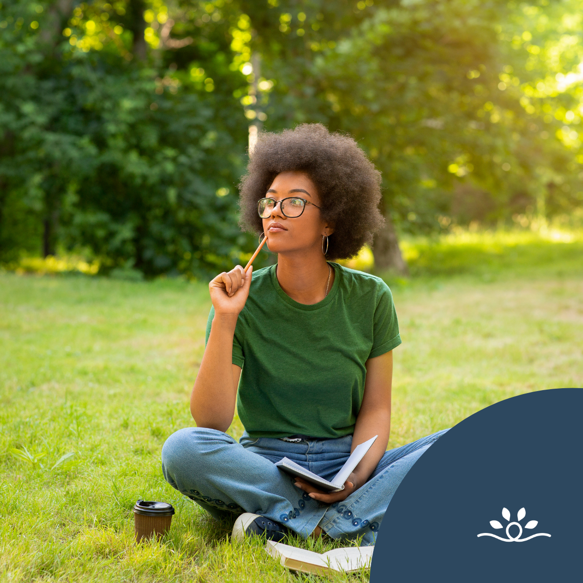 The image depicts a young woman sitting cross-legged on a grassy patch in a park. She has a full, curly afro hairstyle and wears large, round glasses. Dressed in a green t-shirt and light blue jeans, she appears to be deep in thought, holding a pencil to her chin. In her other hand, she holds an open notebook, suggesting she is engaged in some form of writing or contemplation. A small, disposable coffee cup is placed on the ground beside her. The background features lush greenery, with soft sunlight filtering through the trees, creating a serene and inviting atmosphere.