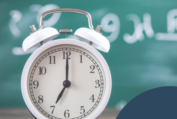 The image features a classic white alarm clock positioned prominently in the foreground. The clock face displays the time just after twelve, with black hour and minute hands set against a cream-colored background. The clock has a round shape with an outer rim and a textured surface, contrasted by two small metal bells at the top. Behind the clock is a blurred green chalkboard, which contains faint, handwritten mathematical symbols and numbers. The background is out of focus, directing attention to the clock. In the upper left corner of the image, there are a series of white dots arranged in a grid pattern. The lower-right corner features a dark blue semicircular shape with a graphic resembling a flower or waves.