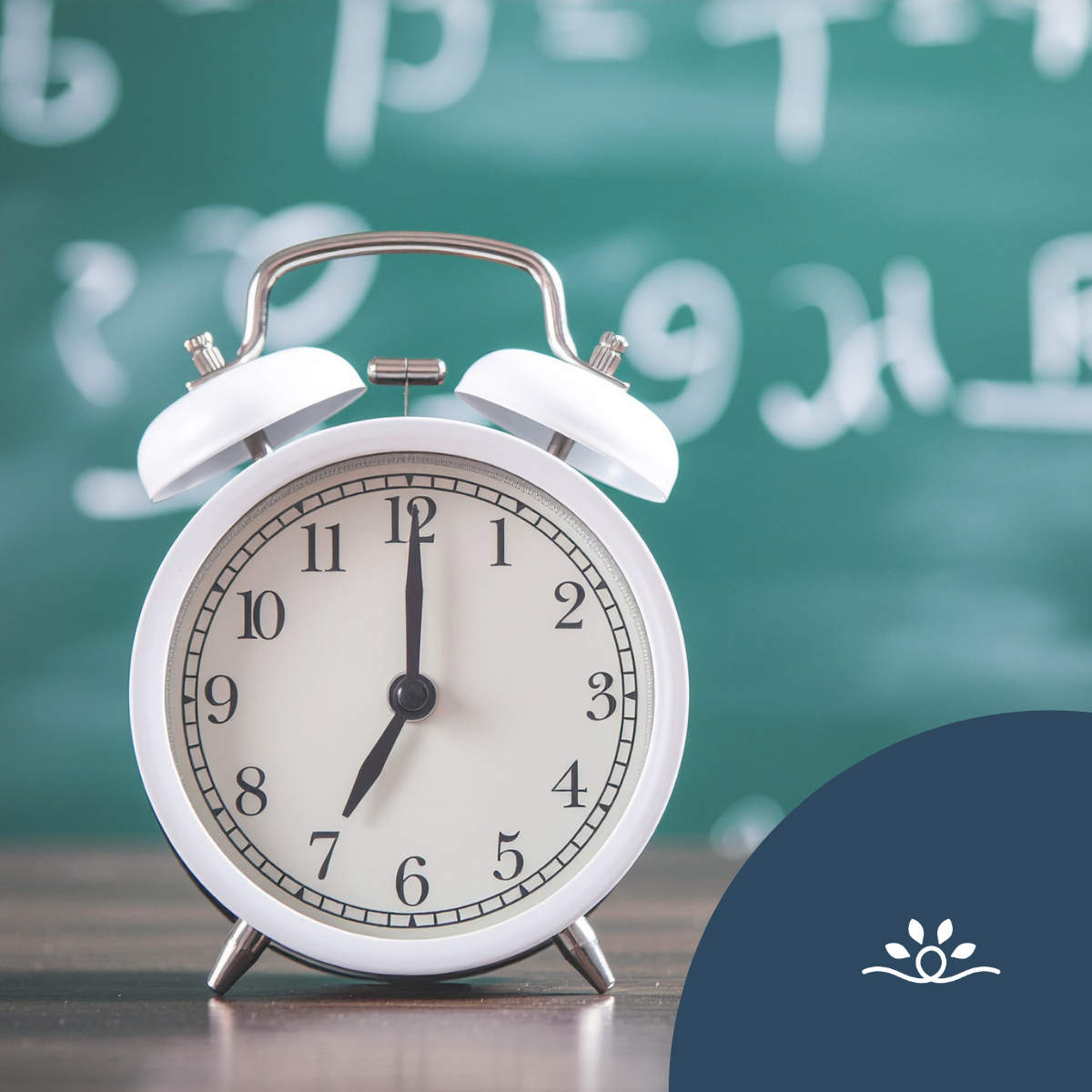 The image features a classic white alarm clock positioned prominently in the foreground. The clock face displays the time just after twelve, with black hour and minute hands set against a cream-colored background. The clock has a round shape with an outer rim and a textured surface, contrasted by two small metal bells at the top. Behind the clock is a blurred green chalkboard, which contains faint, handwritten mathematical symbols and numbers. The background is out of focus, directing attention to the clock. In the upper left corner of the image, there are a series of white dots arranged in a grid pattern. The lower-right corner features a dark blue semicircular shape with a graphic resembling a flower or waves.