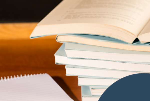 The image features a stack of books placed on a wooden surface, predominantly viewed from the side. The top book is open, showing one page with visible text, while the rest of the books are closed and have pastel blue covers. The colors of the books include soft blue and beige tones, contrasting with the warm brown of the wooden background. In the bottom left corner, there is a spiral-bound notepad, partially visible, with a white cover and lined pages. To the left side of the image, there is a rounded graphic element in a dark blue color, accompanied by a stylized icon of a flower or petal design in white.