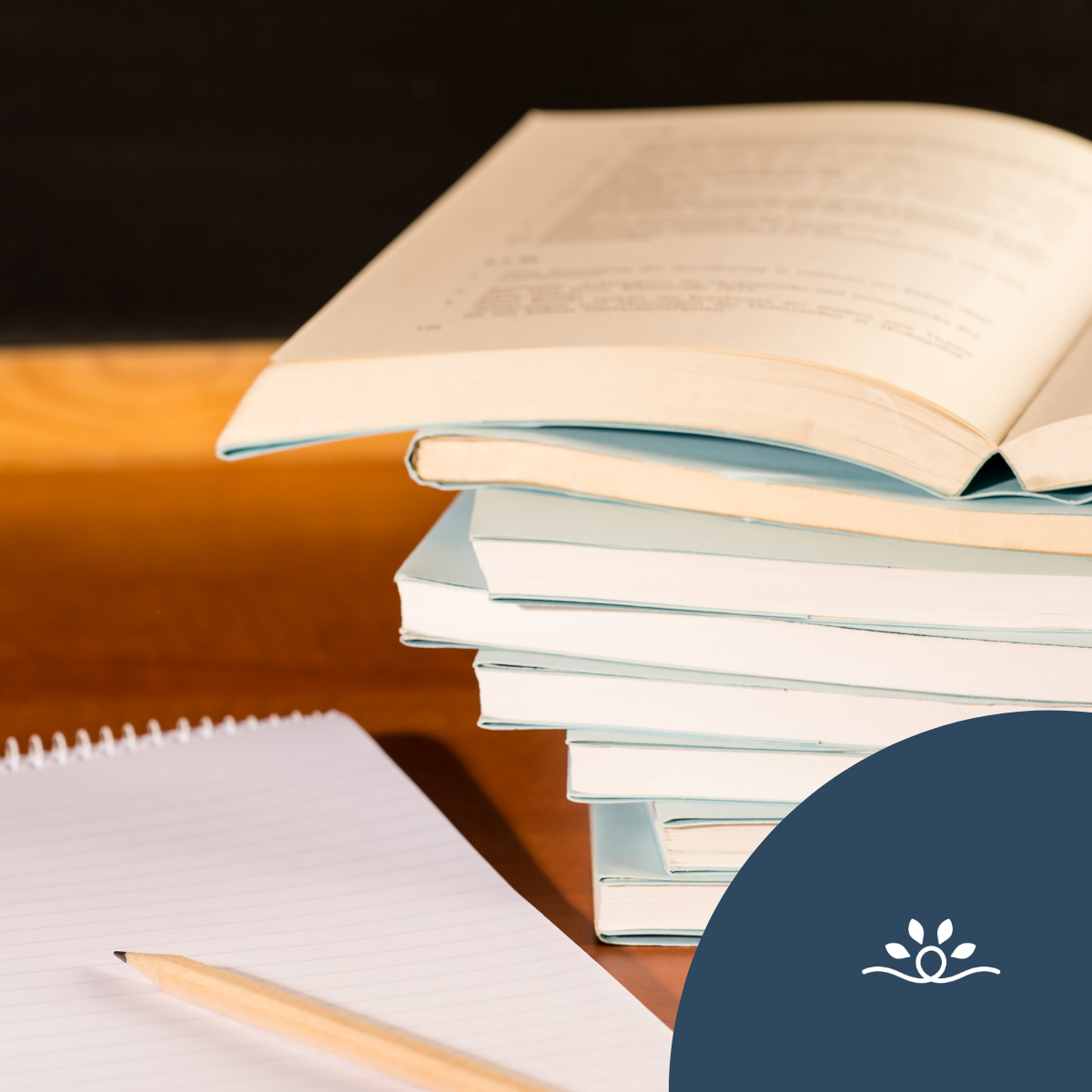 The image features a stack of books placed on a wooden surface, predominantly viewed from the side. The top book is open, showing one page with visible text, while the rest of the books are closed and have pastel blue covers. The colors of the books include soft blue and beige tones, contrasting with the warm brown of the wooden background. In the bottom left corner, there is a spiral-bound notepad, partially visible, with a white cover and lined pages. To the left side of the image, there is a rounded graphic element in a dark blue color, accompanied by a stylized icon of a flower or petal design in white.