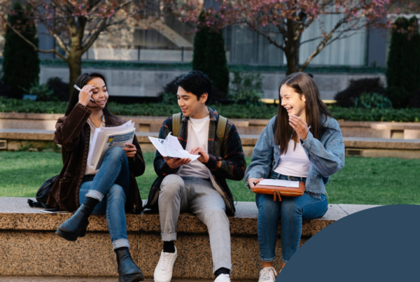 The image depicts three young adults sitting on a stone bench in a green, open area surrounded by trees and shrubs. The scene conveys a casual, collaborative atmosphere with students engaged in studying or discussing their materials. The person on the left is a female, wearing a brown leather jacket and holding multiple sheets of paper, with a pen in hand as she appears to speak. The male in the center is dressed in a white shirt and a plaid jacket; he is facing the girl on the left and is holding a document or book, seemingly listening attentively. The girl on the right, wearing a light blue jacket and holding a notebook, is smiling and appears to be interacting with the others. The background features soft-focus greenery with subtle hints of architecture, contributing to a relaxed outdoor study environment.