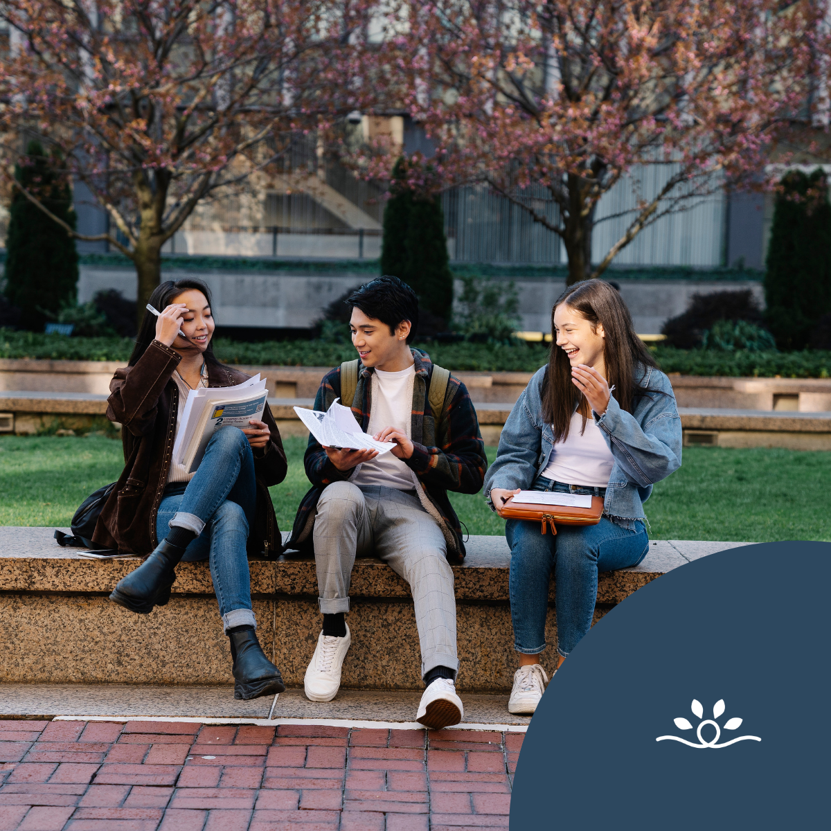 The image depicts three young adults sitting on a stone bench in a green, open area surrounded by trees and shrubs. The scene conveys a casual, collaborative atmosphere with students engaged in studying or discussing their materials. The person on the left is a female, wearing a brown leather jacket and holding multiple sheets of paper, with a pen in hand as she appears to speak. The male in the center is dressed in a white shirt and a plaid jacket; he is facing the girl on the left and is holding a document or book, seemingly listening attentively. The girl on the right, wearing a light blue jacket and holding a notebook, is smiling and appears to be interacting with the others. The background features soft-focus greenery with subtle hints of architecture, contributing to a relaxed outdoor study environment.