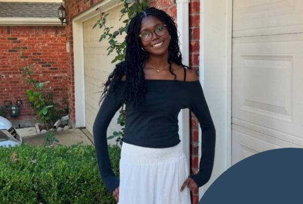 A square outdoor portrait shows a smiling Black woman standing in front of a red-brick house with white garage doors and trim. She wears round eyeglasses, a black off-the-shoulder long-sleeved top, and a long white tiered skirt. Her long black locs fall over one shoulder, and her hands rest near her hips. Green shrubs and climbing vines fill the foreground and grow along the house. A neighboring roof and exterior lantern are visible in the background. A dark blue curved graphic overlay covers the lower-right corner and contains a small white floral or lotus-like symbol.
