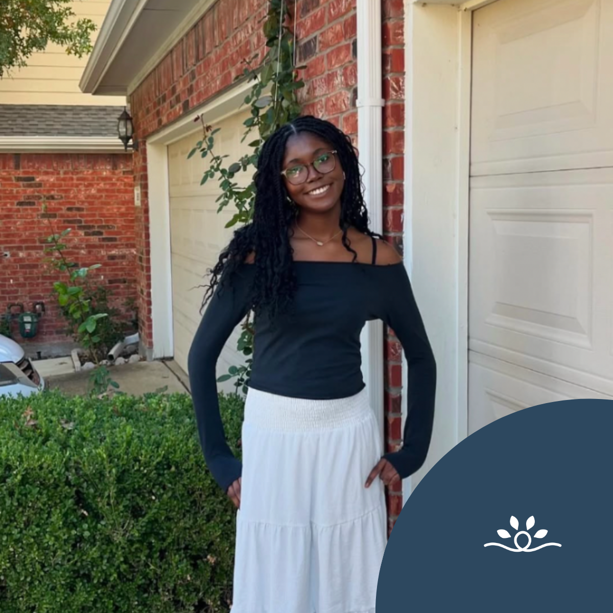 A square outdoor portrait shows a smiling Black woman standing in front of a red-brick house with white garage doors and trim. She wears round eyeglasses, a black off-the-shoulder long-sleeved top, and a long white tiered skirt. Her long black locs fall over one shoulder, and her hands rest near her hips. Green shrubs and climbing vines fill the foreground and grow along the house. A neighboring roof and exterior lantern are visible in the background. A dark blue curved graphic overlay covers the lower-right corner and contains a small white floral or lotus-like symbol.
